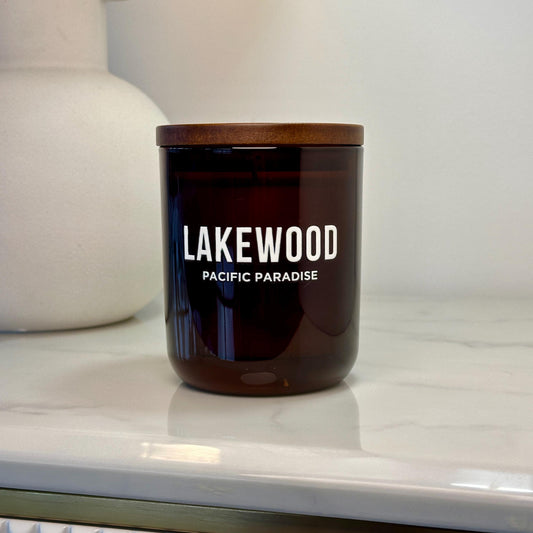 A brown glass candle jar with white “LAKEWOOD” branding and “PACIFIC PARADISE” printed below. The jar is sealed with a wooden lid and sits on a light-colored surface. A white ceramic vase with a soft, rounded shape is in the background.