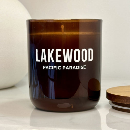 A brown glass candle jar with white “LAKEWOOD” branding and “PACIFIC PARADISE” printed below. The jar sits on a light-colored surface next to a wooden lid. A white ceramic vase with a soft, rounded shape is in the background.