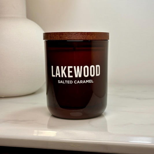 A handcrafted amber glass candle labeled “LAKEWOOD Salted Caramel,” now with its wooden lid on. The candle sits on a white surface against a soft, neutral backdrop, emphasizing its minimalist and elegant design.