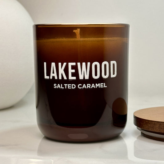 A rich amber glass candle with “LAKEWOOD” and “Salted Caramel” printed in white, placed on a white countertop. The candle is lit, casting a warm glow, with its wooden lid resting beside it. A neutral-toned vase is partially visible in the background.