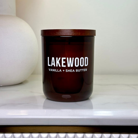 Amber glass Lakewood candle with Vanilla + Shea Butter label and wooden lid, styled against a soft white background on a marble counter.