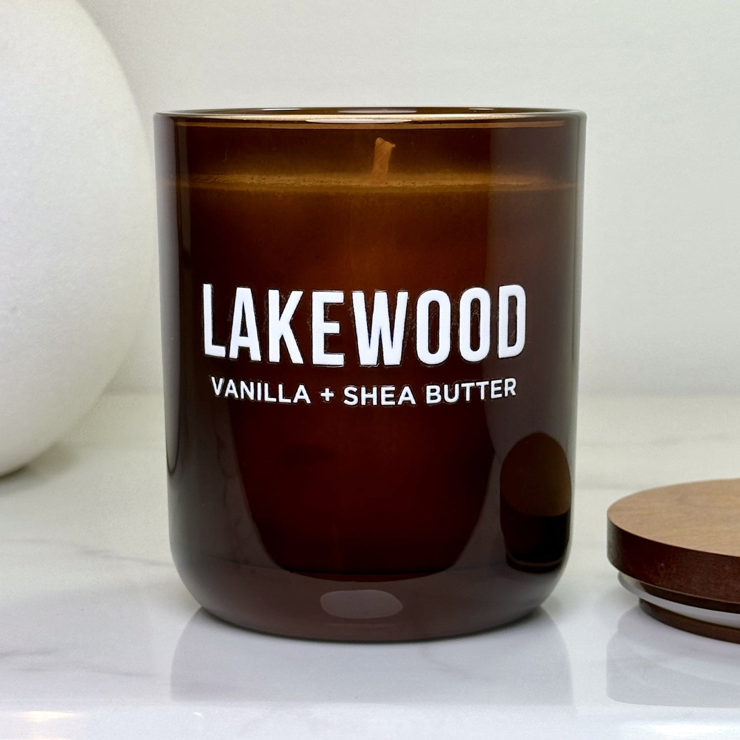 Close-up of the Vanilla + Shea Butter Lakewood candle with lid off, showing the smooth wax and wick inside the amber glass vessel on a marble surface.