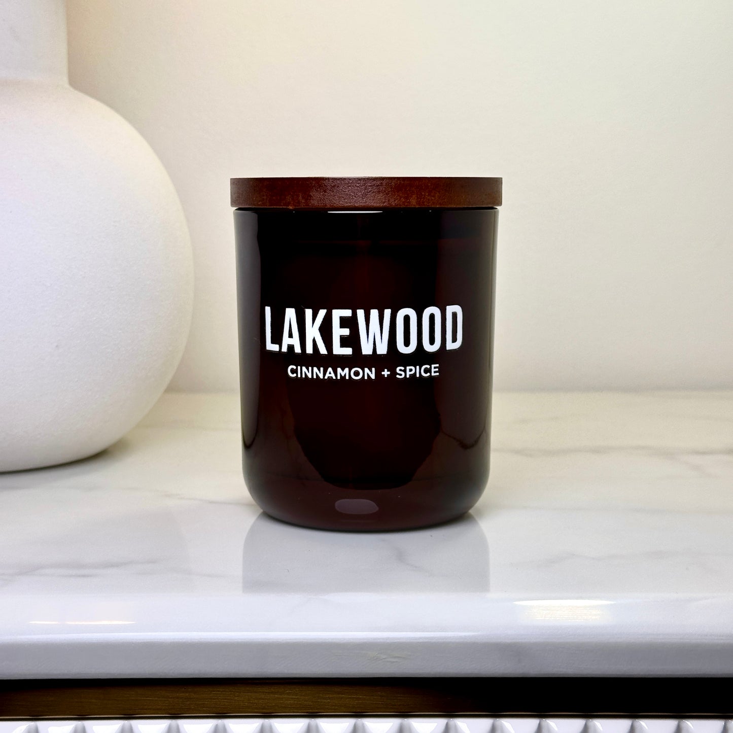 A cinnamon and spice scented candle from Lakewood Candles, featuring a minimalist amber jar with a wooden lid. This handmade soy wax candle delivers a warm, comforting fragrance perfect for cozy spaces. Photographed on a white marble bench with a clean, modern backdrop, it’s an elegant, natural home decor gift. Ideal for thoughtful gifting, autumn vibes, or adding calm to your space. Reusable amber glass jar.
