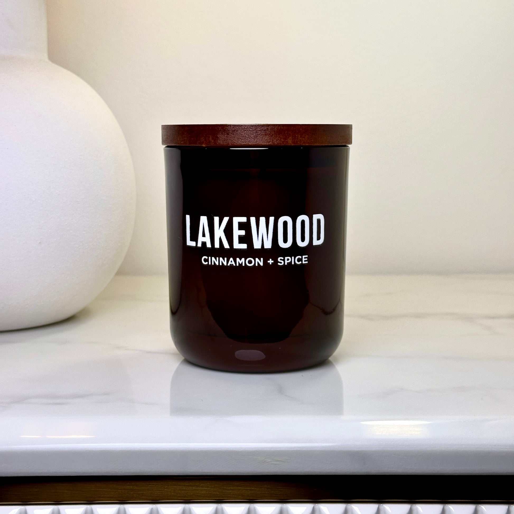 A cinnamon and spice scented candle from Lakewood Candles, featuring a minimalist amber jar with a wooden lid. This handmade soy wax candle delivers a warm, comforting fragrance perfect for cozy spaces. Photographed on a white marble bench with a clean, modern backdrop, it’s an elegant, natural home decor gift. Ideal for thoughtful gifting, autumn vibes, or adding calm to your space. Reusable amber glass jar.